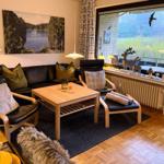 Wohnzimmer mit Sofa, Sessel und Tisch. Große Fensterfront mit Blick auf Grünfläche.