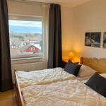 Schlafzimmer mit Blick auf Dachdörfer und Wald. Holzbett, Nachttisch mit Lampe und Bilder an der Wand.