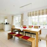 Dining area with wooden table, benches, and chairs. Kitchen in background. Windows with blinds.