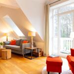 Wohnzimmer mit grauem Sofa, rotem Sessel und Holzboden. Große Fenster mit Blick auf den Garten.