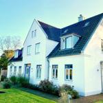 Weiße Villa mit blauen Fensterrahmen und dunklem Dach, umgeben von Grünfläche und Bäumen.