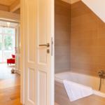 Bad mit Badewanne und Duschbrause. Tür öffnet sich in Wohnbereich mit Holzboden.