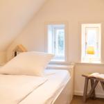 Schlafzimmer mit Bett, zwei Fenstern und Holztisch mit Buch.