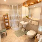 Badezimmer mit Dusche, Waschbecken und Toilette. Holzregal und Handtuchkorb.