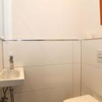 Badezimmer mit Waschbecken, Spiegel und Toilette. Weiße Fliesen und moderne Armaturen.