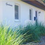 Weiße Hauswand mit Schriftzug, Eingangstür und Fenstern, umgeben von Gräsern und Kies.