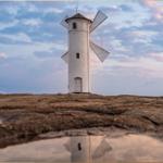 Weißer Windmühlenbau auf felsigem Terrain mit Spiegelung im Wasser.