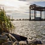 Wasserufer mit Steinen und Schilf, im Hintergrund ein Hafenkran.