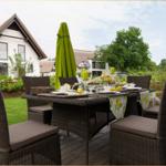 Garten-Terrasse mit gedecktem Esstisch und grünem Sonnenschirm. Hintergrund: weißes Haus mit Dach.