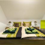 Schlafzimmer mit Doppelbett, grünen Kissen und Stuhl unter schrägem Dach.