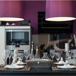 Gedeckter Esstisch mit Gläsern und Kerzen in moderner Küche mit lila Deckenleuchten.