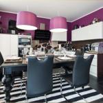 Essbereich mit Tisch, Stühlen und Küchenzeile. Wandfarbe lila, Deckenleuchten und Teppich mit Streifen.