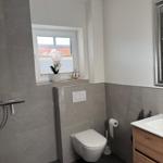 Badezimmer mit Toilette, Waschbecken und Fenster. Graue Fliesen und Holzschrank.