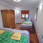 Zwei Betten mit grünem Bettzeug, Holzschrank und Fenster mit rotem Rollladen.