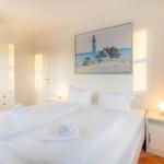 Schlafzimmer mit weißem Bett, Nachttisch und Wandbild mit Leuchtturm.