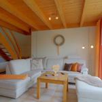 Wohnzimmer mit weißem Sofa, Holztisch und Holzbalkendecke. Große Fensterfront mit orangefarbenen Vorhängen.