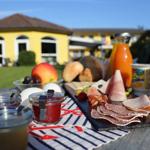 Frühstückstablett mit Wurst, Käse, Obst und Joghurt auf Holztisch vor gelbem Haus