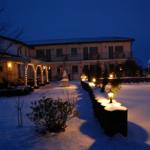 Schneebedeckter Hof mit beleuchteten Lampen und verschneiten Pflanzen vor einem zweistöckigen Haus.