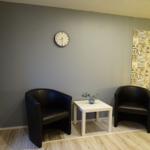 Zwei schwarze Sessel und ein weißer Tisch mit Blumen vor einer Wand mit Uhr und Wandbild.