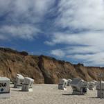 Strand mit weißen Strandkörben und sandigem Ufer vor steilen Klippen.