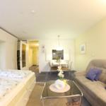 Wohnung mit Schlafzimmer, Sofa und Essbereich. Glas-Tisch mit Blumen.