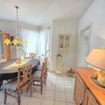 Dining room with wooden table, chairs, and lighting. Background: windows, cabinets, and lamps.
