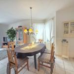 Dining room with round table, wooden chairs, and chandelier. Windows with white curtains.