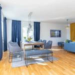 Esszimmer mit Holztisch, grauen Stühlen und blauen Vorhängen. Wohnbereich mit blauem Sofa und gelben Vorhängen.