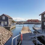 Holzhäuser am Wasser mit Steg und rotem Bootshaus im Hintergrund.