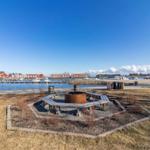 Feuerstelle mit Sitzbank am Wasser, Hintergrund mit Booten und roten Häusern.
