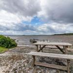 Holztisch und Bänke auf Steinufer mit Blick auf See und Himmel.