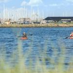 Zwei Personen paddeln in orangefarbenen Kanus auf dem Wasser vor einem Hafen mit Booten.