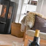Ein Kamin mit brennendem Holz, ein Sessel mit Felldecke und Kissen, ein Korb und ein Flaschenhals im Vordergrund.