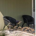 Zwei dunkle Rattan-Stühle stehen auf einer Terrasse vor einer grünen Holzwand.