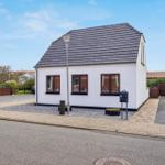 A white house with brown windows and a gray roof stands on a street.