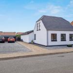 White house with dark roof, paved path, and gravel area. Two cars parked in front of the house.