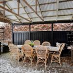 Outdoor area with table and chairs under roof, snow on floor and hedges.