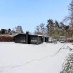 Black wooden house with snow-covered ground and trees in the background.