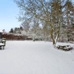 Snow-covered outdoor area with wooden terrace, table, and trees.