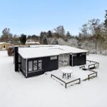 Black-painted cabin with snow-covered roof and terrace in winter.