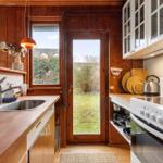 Küche mit Holzwand, Fenster und Tür zum Garten. Weiße Schränke, Edelstahlkochtopf und Arbeitsfläche.