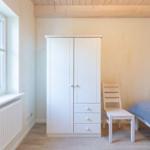 Schlafzimmer mit Bett, Kleiderschrank und Stuhl. Holzdecke und Fenster mit Heizkörper.