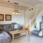 Wohnzimmer mit Sofa, Sessel und Holztisch. Hintergrund: Treppe und Kaminofen.