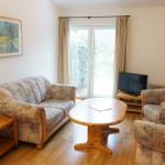 Wohnzimmer mit Sofa, Sesseln, Esstisch und TV. Fenster mit Vorhängen.