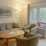 Wohnzimmer mit Sofa, zwei Sesseln und Holztisch. Fenster mit Blick auf Garten.