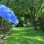 Garten mit Hängemöbel unter blauem Sonnenschirm, Holzbank und Bäumen.