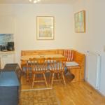 Essbereich mit Holztisch, Bänken und Stühlen. Küche und Kachelofen sichtbar.