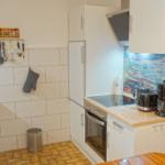 Küche mit weißen Schränken, Herd und Kühlschrank. Arbeitsfläche aus Holz. Wand mit Bild und Küchenutensilien.