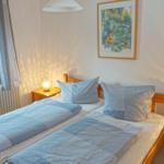 Zwei Einzelbetten mit blauen Bettwäschen und Holzrahmen. Fenster mit Vorhängen und Wandbild.