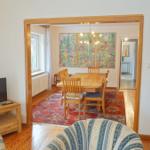 Wohnzimmer mit Fernseher und Sitzgelegenheiten. Durchgang zu Esszimmer mit Tisch, Stühlen und Wandbildern.